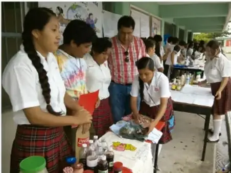 Fotografía de estudiantes de secundaria en uniforme organizando y atendiendo un puesto del Festival literario, con materiales y juegos sobre una mesa, en el patio escolar.