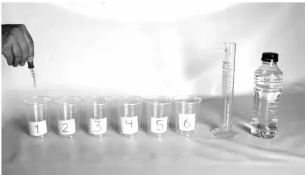 Fotografía de seis vasos plásticos alineados y rotulados con los números 1 al 6, una probeta graduada con agua, una botella plástica y una mano sosteniendo un gotario sobre el primer vaso.