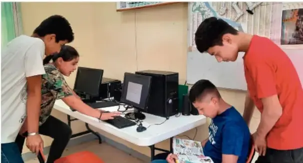 Fotografía de cuatro estudiantes (tres jóvenes de pie y una sentada) reunidos alrededor de una mesa con dos computadoras y papeles, trabajando en equipo en un aula. Crédito: Jesús Eduardo Arteaga Flores, Tamaulipas.
