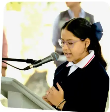 Niña con lentes y uniforme escolar habla frente a un micrófono, posiblemente en una asamblea escolar.
