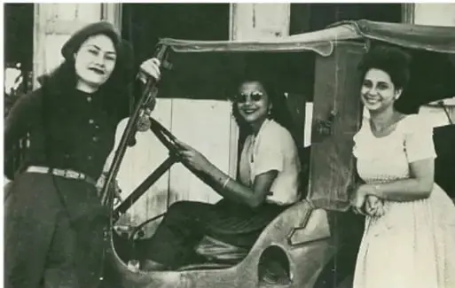 Fotografía en blanco y negro de las hermanas Mirabal junto a un automóvil descapotable: una de pie al volante y las otras dos sonrientes en el vehículo.