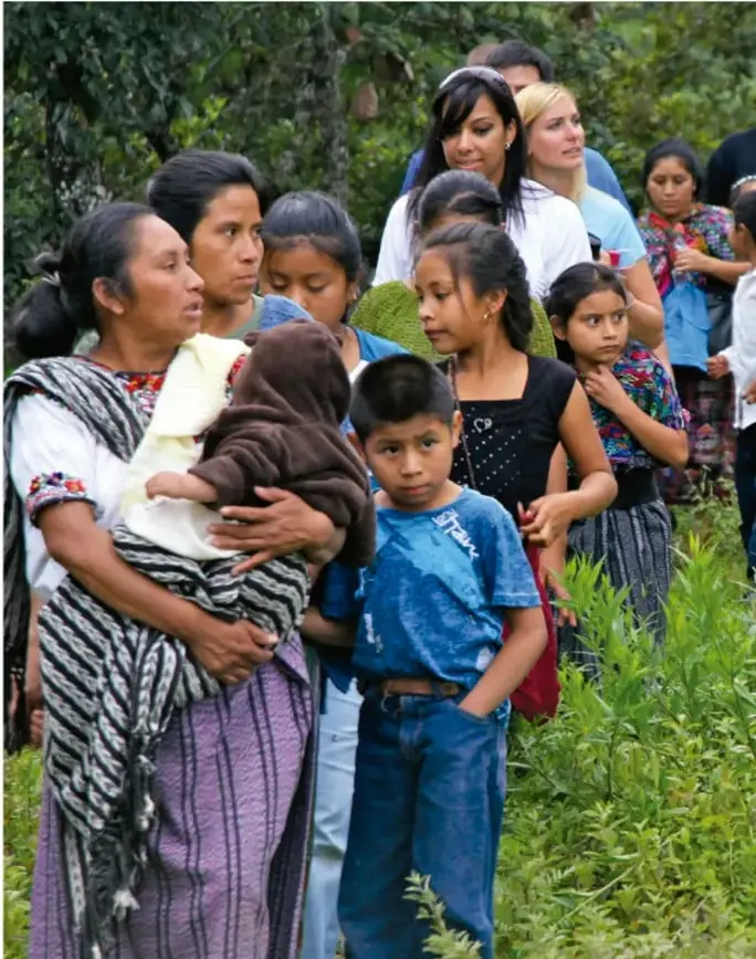 Fotografía de una comunidad indígena en Chiapas: mujeres con.trajes tradicionales, niñas y niños reunidos al aire libre entre vegetación, posiblemente durante una asamblea o evento comunitario.
