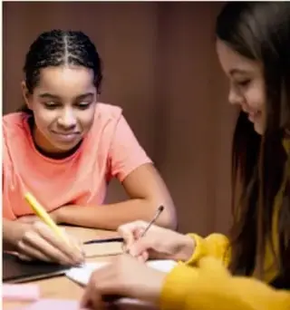 Fotografía de dos estudiantes trabajando juntas en una mesa, una escribe en un cuaderno mientras la otra observa; ilustra el trabajo colaborativo del proyecto.