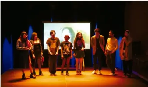 Fotografía de un grupo de estudiantes de telesecundaria de pie en un escenario iluminado, frente a una pantalla de proyección, realizando una presentación o dramatización ante su comunidad escolar.