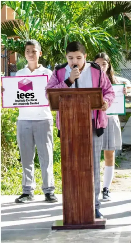 Tres estudiantes de Telesecundaria en un acto cívico al aire libre: uno habla en un atril de madera con micrófono y otros dos sostienen carteles, uno con el logo del Instituto Electoral del Estado de Sonora (iees).