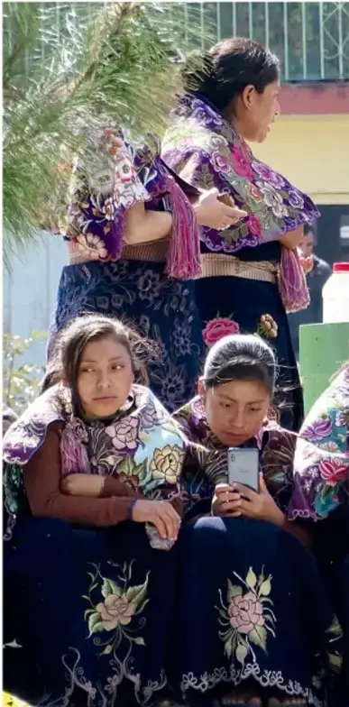 Fotografía de jóvenes mujeres con blusas y faldas bordadas de flores en tonos morados y negros, sentadas; una de ellas mira la pantalla de un teléfono celular; al fondo hay ramas de árbol.