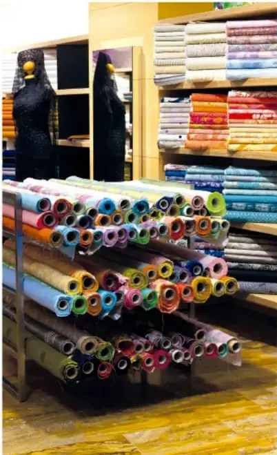 Fotografía de una tienda de telas con estantes llenos de rollos de tela de colores. Al fondo se ven dos maniquíes con vestidos negros. La imagen ilustra el consumo virtual de agua, ya que la producción de telas requiere grandes cantidades de agua.