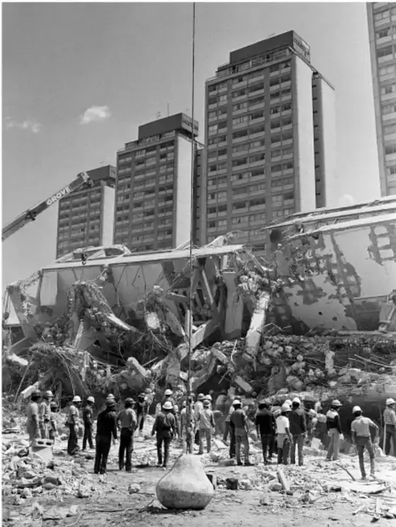 Fotografía en blanco y negro de un edificio derrumbado tras el sismo de 1985 en la Ciudad de México. Se observan escombros, estructuras colapsadas y un grupo de personas, algunas con cascos, entre los restos. Al fondo, edificios de departamentos aún en pie.