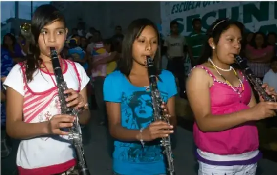 Fotografía de tres estudiantes tocando clarinetes en un evento escolar, con público y la leyenda ESCUE... SECUN... de fondo.