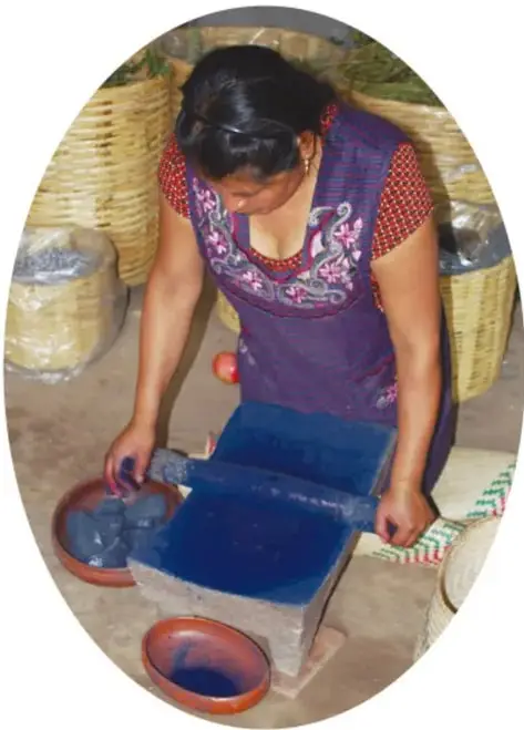 Fotografía de una mujer teñiendo telas con añil: sostiene una tela empapada en un azul profundo, junto a recipientes con el tinte y cestos, en un taller artesanal.