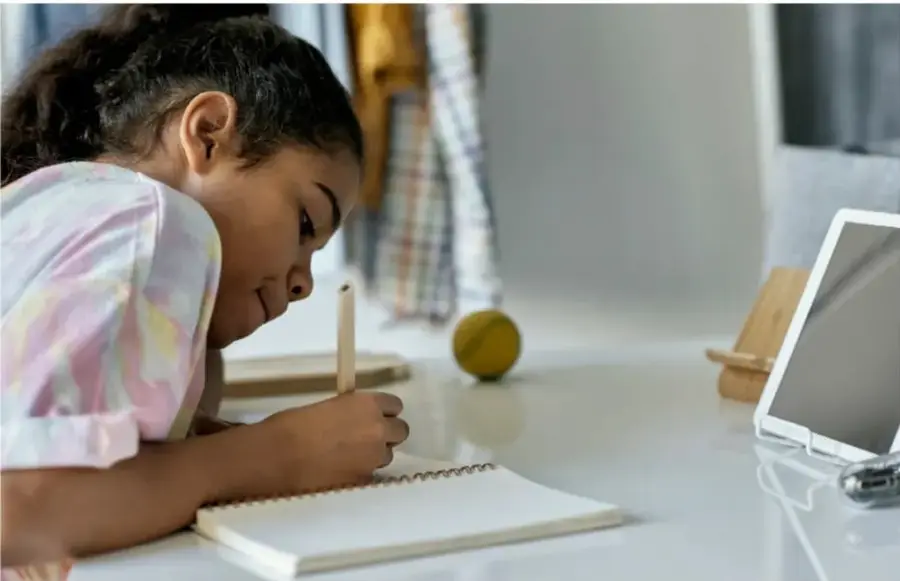 Niña de primaria escribiendo en un cuaderno sobre un escritorio blanco, con una tableta digital y una pelota de tenis al fondo.