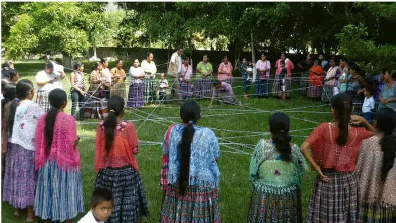 Fotografía de un grupo de personas (comunidad) formando un círculo al aire libre en un patio con árboles, sosteniendo cuerdas que se cruzan en el centro, ilustrando una actividad lúdica colectiva.