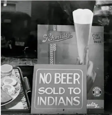 Fotografía histórica en la que se ve un letrero en un establecimiento que dice 'NO BEER SOLD TO INDIANS' (No se vende cerveza a indios), junto a publicidad de cerveza Schmidt's; ilustra un caso de discriminación racial por señalamiento.