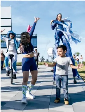 Fotografía de jóvenes y un niño en una plaza pública; una joven en zancos con vestimenta azul ofrece un espectáculo mientras un niño la mira sonriente; sugiere la campaña artística en espacios públicos.