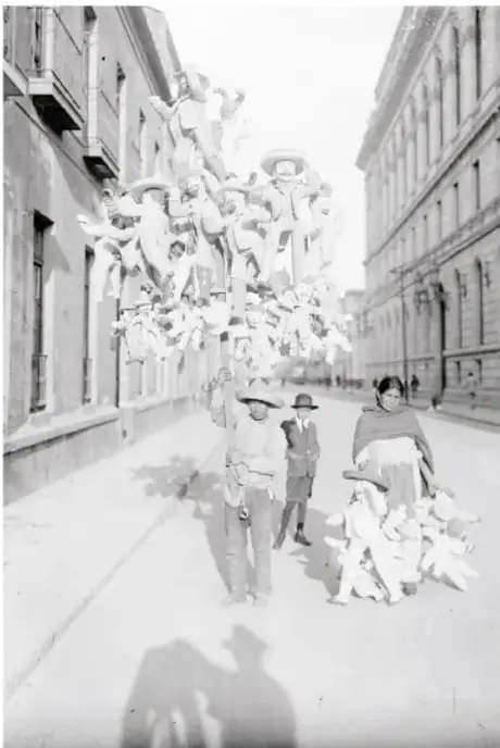 Fotografía en blanco y negro de personas que cargan grandes ramos de globos con figuras (almidón o juguetes) mientras caminan por una calle con edificios clásicos; un adulto y niños los observan.