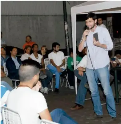 Fotografía de una reunión comunitaria: un hombre de pie con micrófono habla ante un grupo de personas sentadas, algunas con playeras verdes, en un espacio techado.