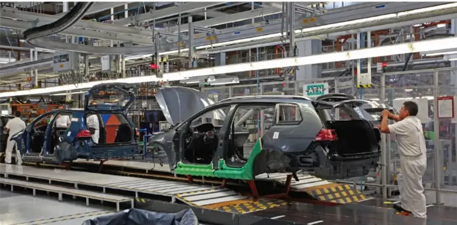 Planta automotriz con una línea de ensamblaje: varios automóviles en distintas etapas de armado, operarios con uniforme y maquinaria industrial.