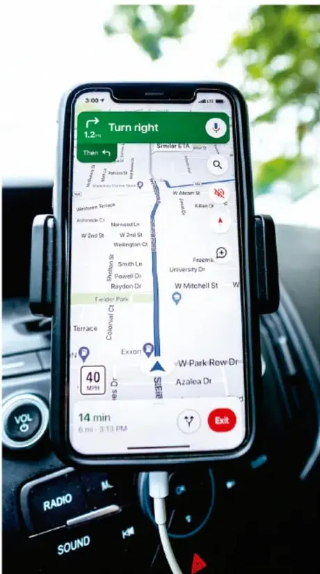 Fotografía de un teléfono inteligente sostenido en un soporte dentro de un automóvil, mostrando una aplicación de navegación GPS con indicaciones de ruta ("Turn right"), velocidad de 40 MPH y tiempo estimado de llegada de 14 min.
