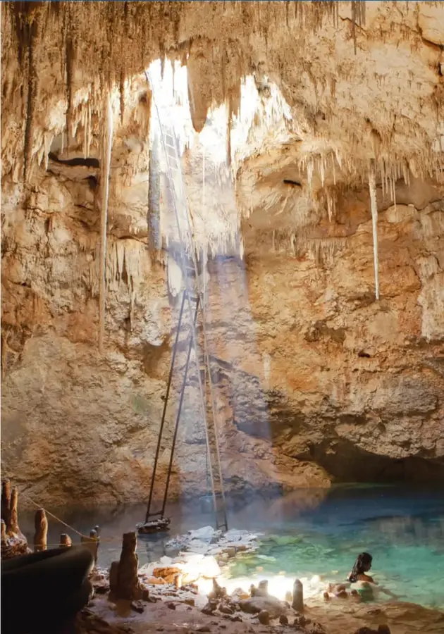 Fotografía de un cenote: gran abertura en la roca caliza con estalactitas colgantes, un haz de luz solar que entra por una claraboya, una escalera de madera apoyada en el agua turquesa y una persona bañándose; al fondo, una embarcación pequeña. La imagen evoca la relación entre naturaleza, territorio y sociedad.