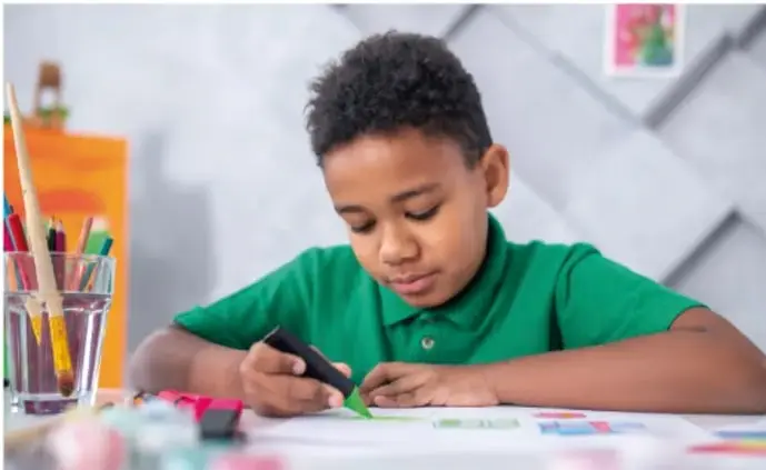 Fotografía de un niño escribiendo o dibujando en una hoja, con colores y lápices sobre la mesa.