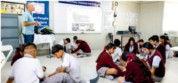 Fotografía de un aula donde estudiantes con uniforme trabajan sentados en el piso en equipos, mientras un maestro observa junto a un proyector y una pizarra.