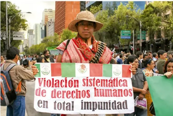 Fotografía de una manifestación en una avenida de una ciudad mexicana; en primer plano una mujer con sombrero de paja y traje regional sostiene una pancarta que dice "Violación sistemática de derechos humanos en total impunidad"; alrededor hay manifestantes con mochilas y más pancartas; se ven árboles y edificios al fondo.
