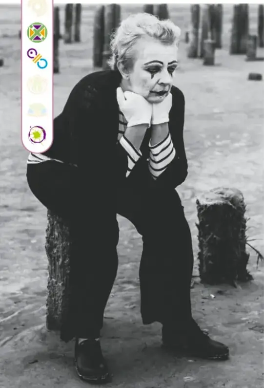 Fotografía en blanco y negro de una persona maquillada como mimo, sentada y pensativa, con vestuario de rayas y maquillaje teatral.