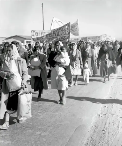 Fotografía en blanco y negro de un grupo de personas caminando por un camino de tierra. Se observan mujeres con cobijas, niños y hombres con pancartas. Representa la Caravana del hambre de los mineros de Coahuila hacia la Ciudad de México en 1950.