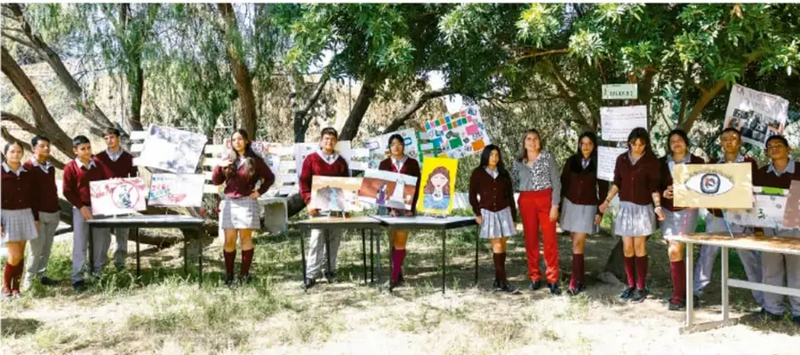 Fotografía de estudiantes de telesecundaria con uniforme presentando afiches y expresiones artísticas en mesas al aire libre, en un patio con árboles, ante la comunidad escolar.
