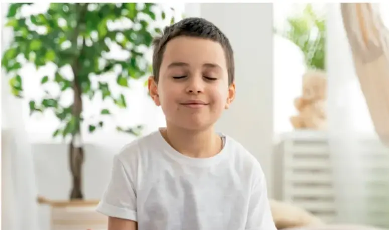 Niño con los ojos cerrados y expresión tranquila, con las manos cerca del abdomen, practicando la respiración consciente en un ambiente con plantas.