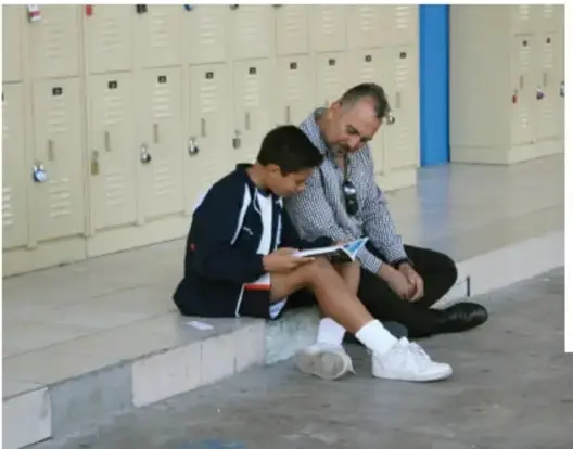 Un adulto y un niño sentados en el piso frente a unos casilleros, leyendo o trabajando juntos en un cuaderno.