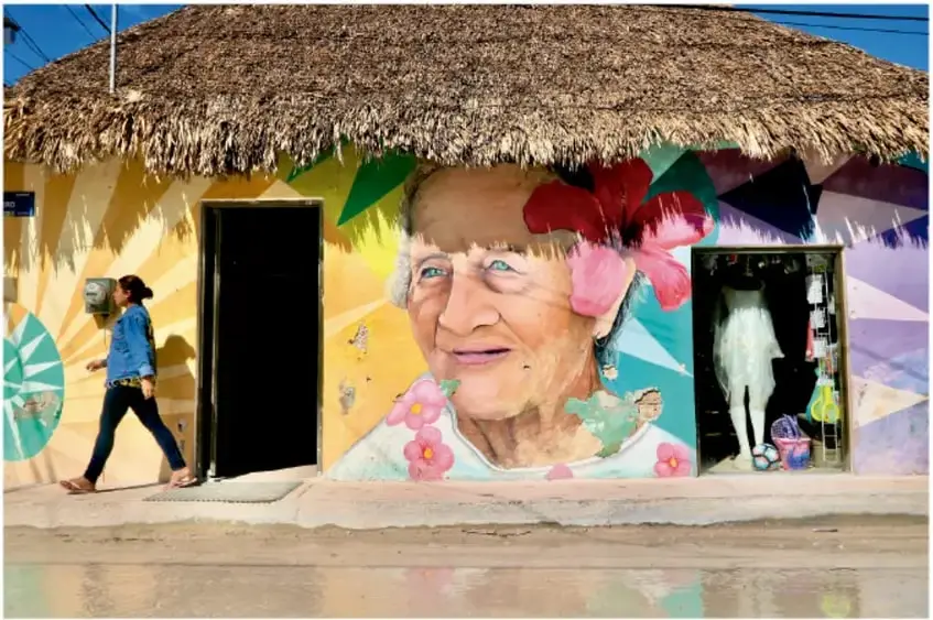 Fotografía de una casa con techo de palapa y mural que retrata a una mujer mayor, en Holbox, Quintana Roo; una persona camina por la banqueta.