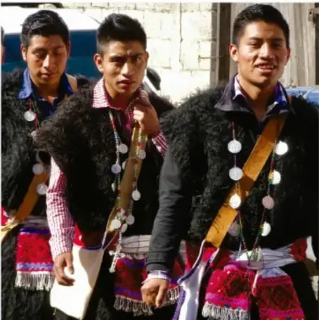 Fotografía de tres jóvenes de una comunidad indígena vistiendo ponchos oscuros tradicionales adornados con monedas, cintas y franjas de colores rojo, azul y verde, junto a un muro de ladrillo.