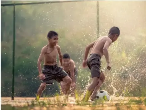 Fotografía de tres niños descalzos jugando con un balón entre salpicaduras de agua en la hierba, al aire libre.
