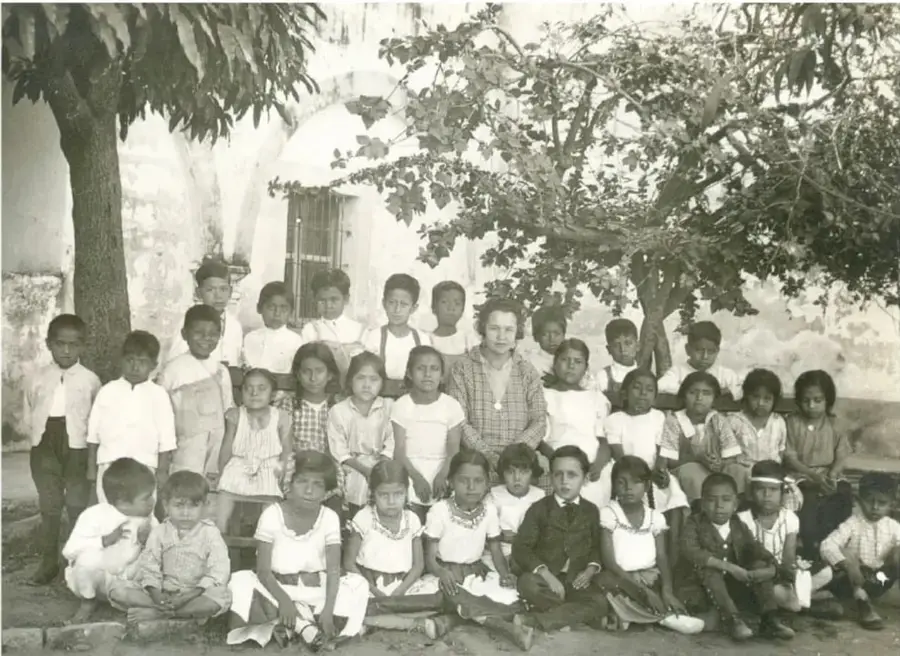 Fotografía antigua en blanco y negro de un grupo numeroso de niños y una maestra frente a un edificio con un árbol grande.