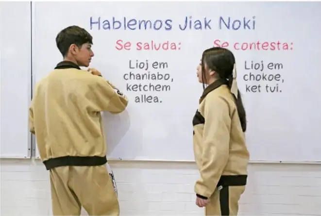 Fotografía de dos estudiantes de secundaria frente a una pizarra con el encabezado 'Hablemos Jiak Noki'. Indica la forma de saludar ('Lioj em chaniabo, ketchem alleya') y la de contestar ('Lioj em chokoe, ket tul') en una lengua originaria.