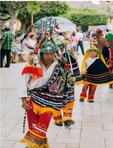 Fotografía de una celebración comunitaria al aire libre con danzantes de trajes tradicionales coloridos, niños y público observando.