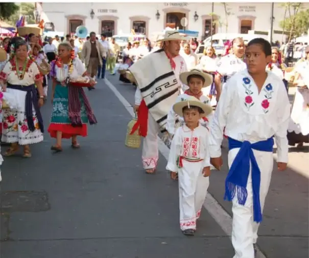Fotografía de un desfile o callejoneada donde niños, jóvenes y adultos visten trajes típicos regionales de México (blusas bordadas, rebozos, sombreros, fajas azules) caminando por una calle con edificios coloniales al fondo.