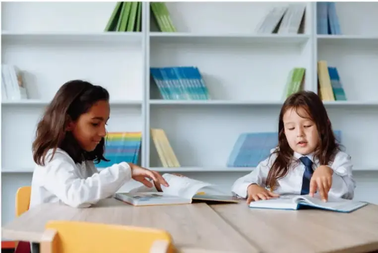 Dos niñas sentadas en una mesa de la biblioteca escolar hojeando un libro; al fondo, estanterías con libros.