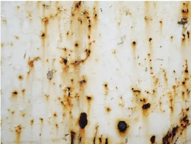Fotografía de una superficie metálica blanca con manchas de óxido color café y negras, ilustrando la corrosión del hierro.
