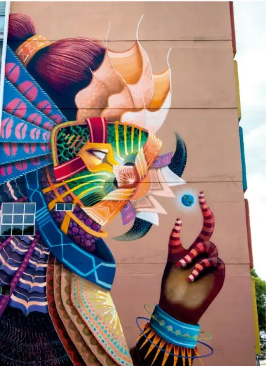 Mural o pintura muralista de gran colorido que muestra una figura con rasgos indígenas, penacho de plumas y un sostenido de colores vivos, con patrones geométricos y vegetales, ilustrando los recursos artísticos visuales mencionados en el texto.