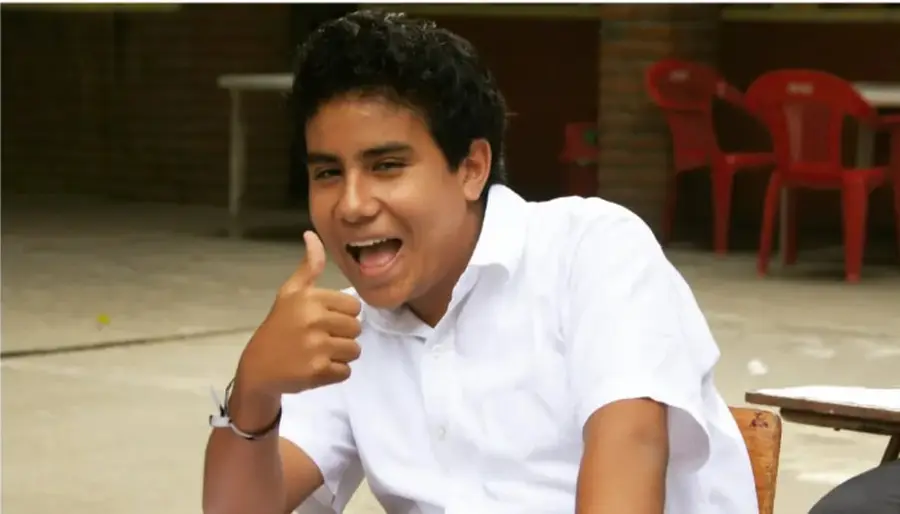 Fotografía de un joven con camisa blanca dando el pulgar arriba y sonriendo, sentado en un patio escolar con sillas rojas y mesas de ladrillo al fondo.