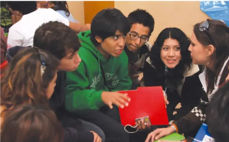 Fotografía de un grupo de adolescentes sentados en círculo en el piso, trabajando en equipo alrededor de un cuaderno rojo; una de ellas habla y los demás escuchan atentamente.