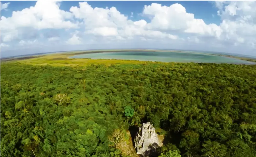 Fotografía aérea de un paisaje natural: una extensa selva verde con un cuerpo de agua (laguna o mar) al fondo, bajo un cielo con nubes. En primer plano se observa una estructura rocosa o edificio antiguo entre la vegetación. La imagen ilustra la relación entre naturaleza y cultura.