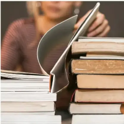 Fotografía de una persona leyendo un libro abierto, apoyado sobre una pila de libros.