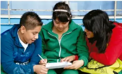 Fotografía de tres estudiantes (dos niñas y un niño) sentados juntos revisando un cuaderno, posiblemente preparando el proyecto escolar.