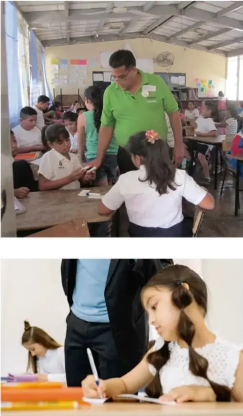 Fotografía de un aula con un maestro de camisa verde revisando el trabajo de una estudiante, mientras otros alumnos trabajan en sus mesas.