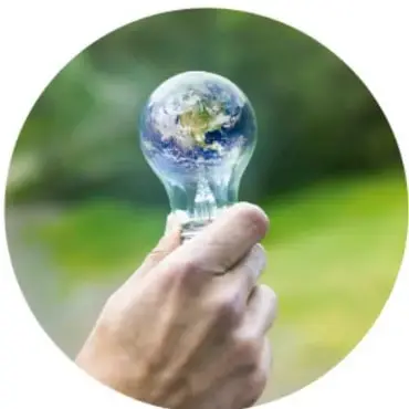 Mano sosteniendo una bombilla cuyo interior muestra el planeta Tierra, símbolo de iluminación limpia y energía sustentable sobre fondo verde difuso.