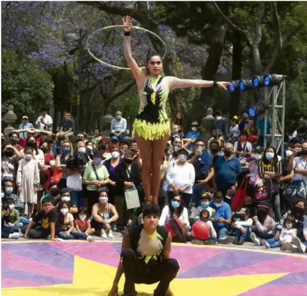 Fotografía de teatro callejero en un parque: una acróbata con malla negra y amarilla equilibra un aro en la mano mientras está de pie sobre otra persona en cuclillas, sobre una lona morada y amarilla, ante un numeroso público con cubrebocas.