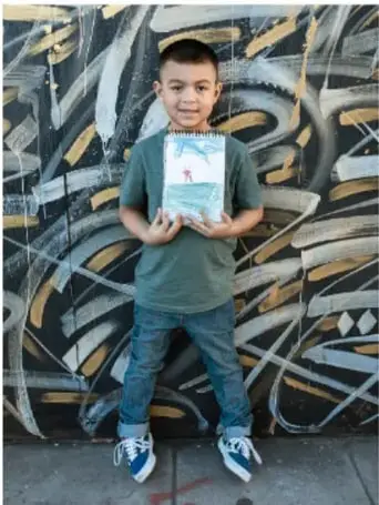 Fotografía de un niño sosteniendo una libreta decorada frente a un mural.
